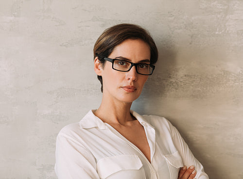 Portrait of a middle-aged businesswoman in eyeglasses leaning on a wall looking straight at a camera