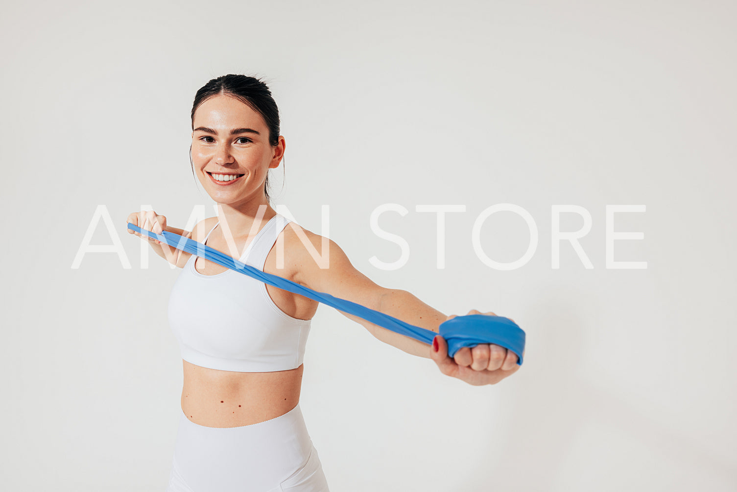 Cheerful woman in white fitness wear is warming up her arms with a resistance band in the studio