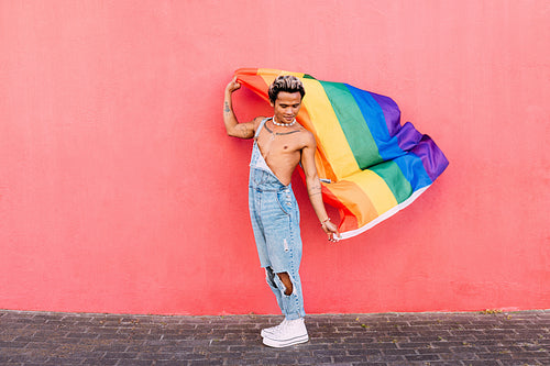 Gay male holding a rainbow LGBT flag in city against pink wall