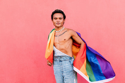 Young man wrapped in rainbow LGBT flag standing outdoors and looking at camera