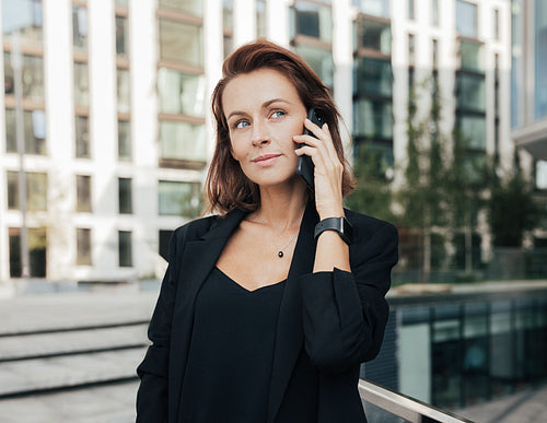 Portrait of a middle-aged female looking away while taking on a mobile phone. Corporate person with ginger hair standing outdoors and having a conversation by smartphone.