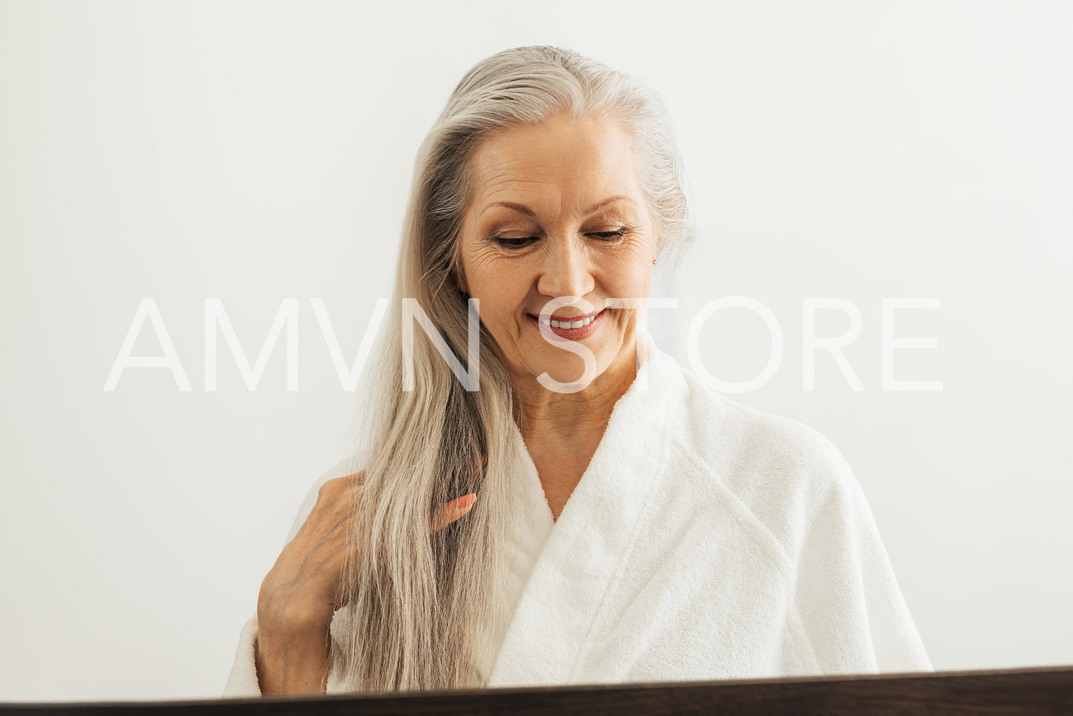 Senior woman in bathrobe looking down while adjusting her long white hair while in front of a mirror