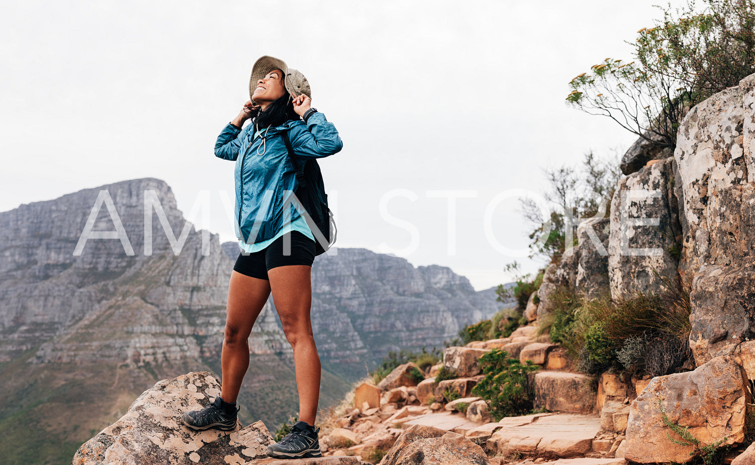 Happy woman hiker holding her hat while looking up standing on a top of a mountain