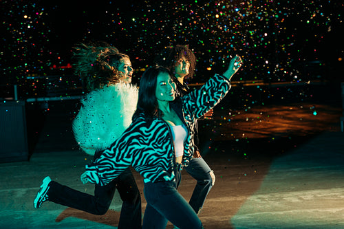 Happy female friends running together on the roof and throwing confetti at night. Three stylish girls celebrating outdoors.