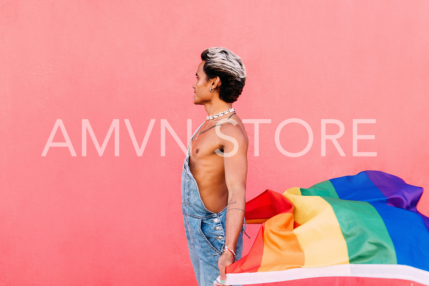 Side view of young stylish guy holding a rainbow LGBT flag against pink wall