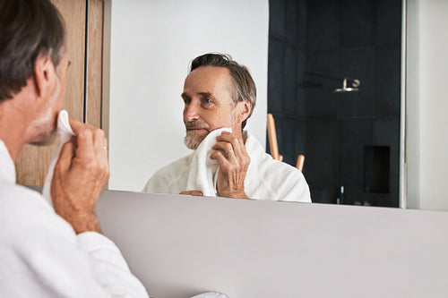 Handsome mature man wiping his cheek with a towel in a bathroom in front of a mirror