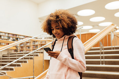 Girl with smartphone in library. Young female with backpack on stairs in library.