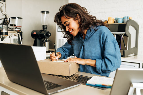 Young smiling entrepreneur packaging order in coffee shop. Woman writing on cardboard box.