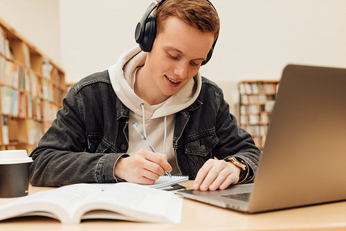 Male student preparing exams at desk in library. Smiling guy in headphones writing on notebook.