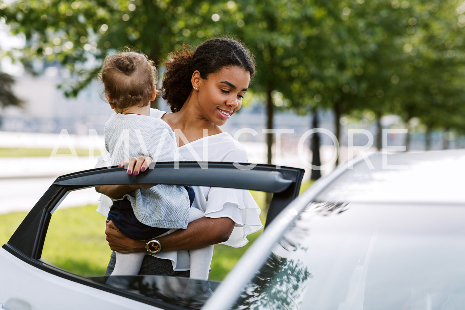 Woman opening a car door and putting her little daughter into it. Young female holding little girl on hands near a car.	