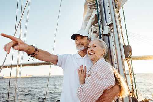 Mature man pointing into the distance and hugging his wife. Two smiling people enjoying a boat trip at sunset.