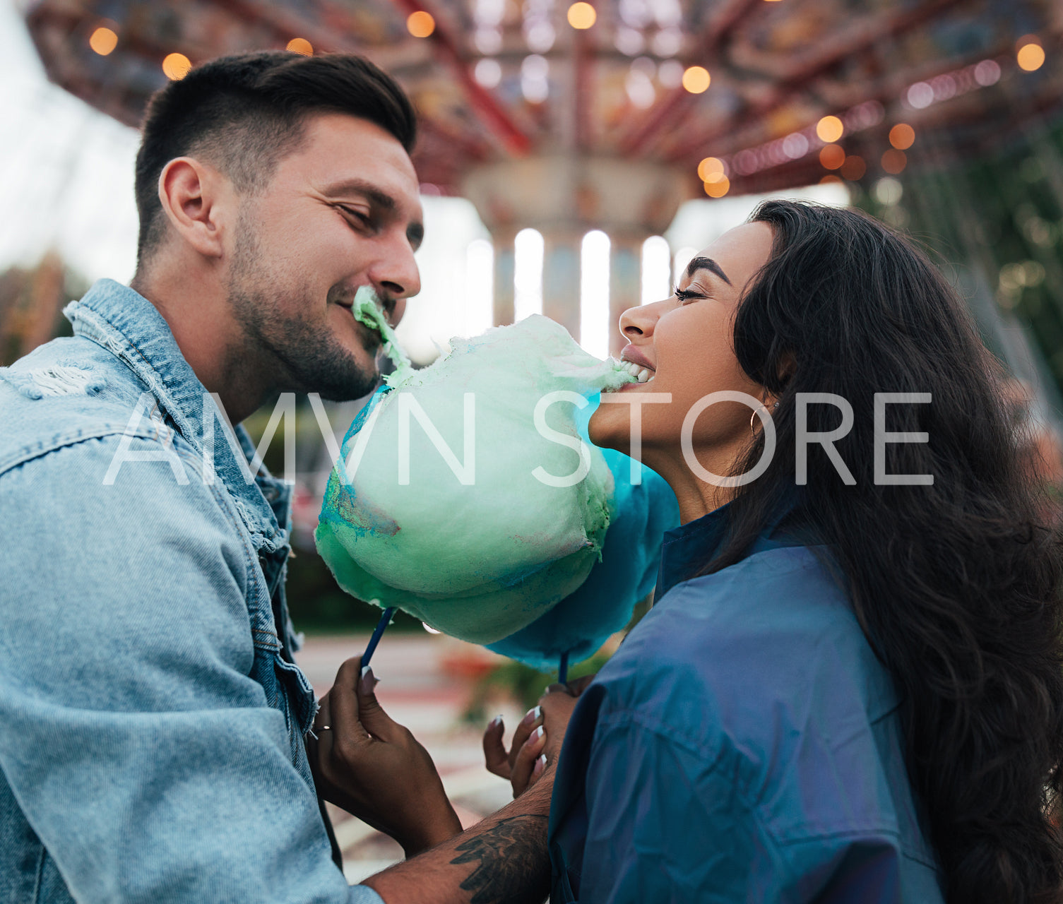 Young happy couple biting cotton candy. Man and his girlfriend bite a blue cotton candy against the carousel.