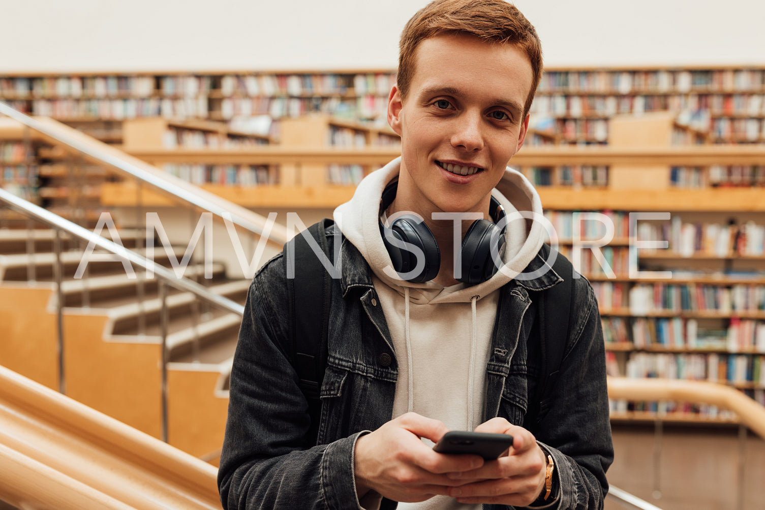 Portrait of a handsome student in casuals standing in library with smartphone