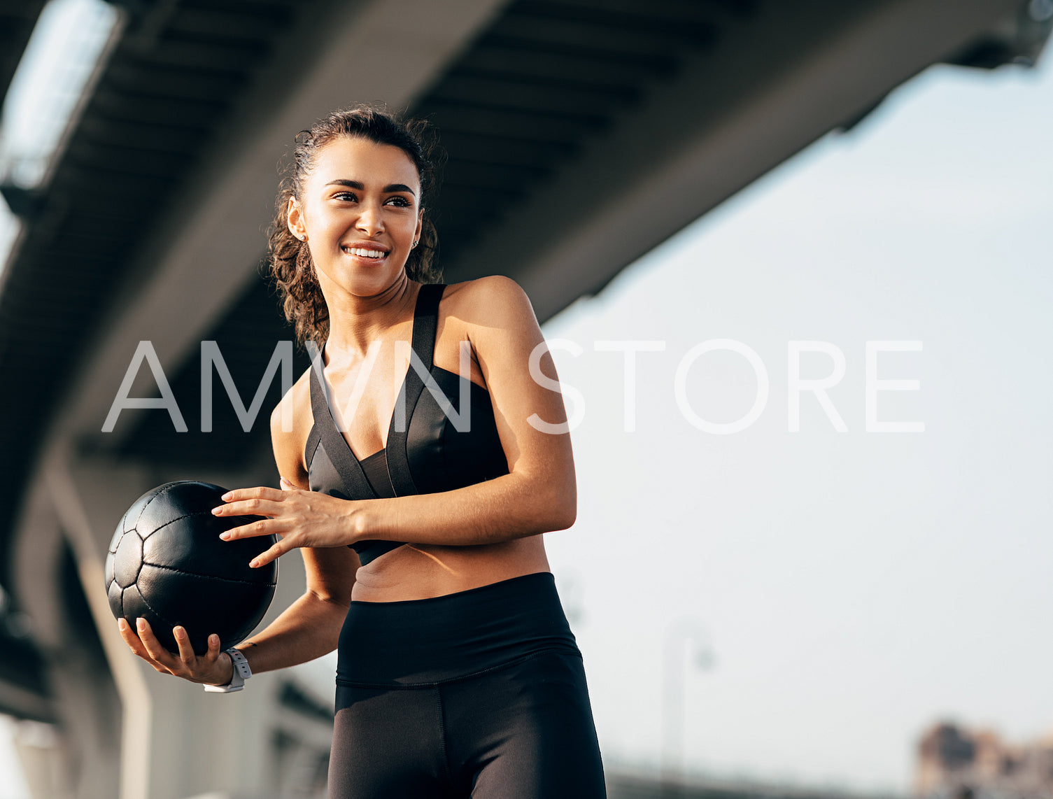 Smiling fit woman holding a medicine ball under a highway and looking away	