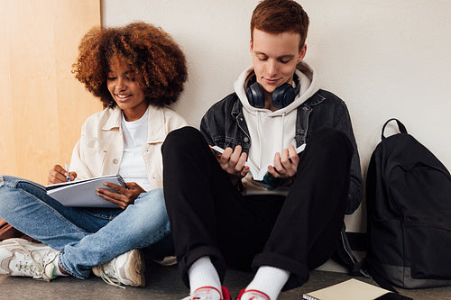 Two classmates sitting at wall in school. Students preparing their assignment while sitting together.