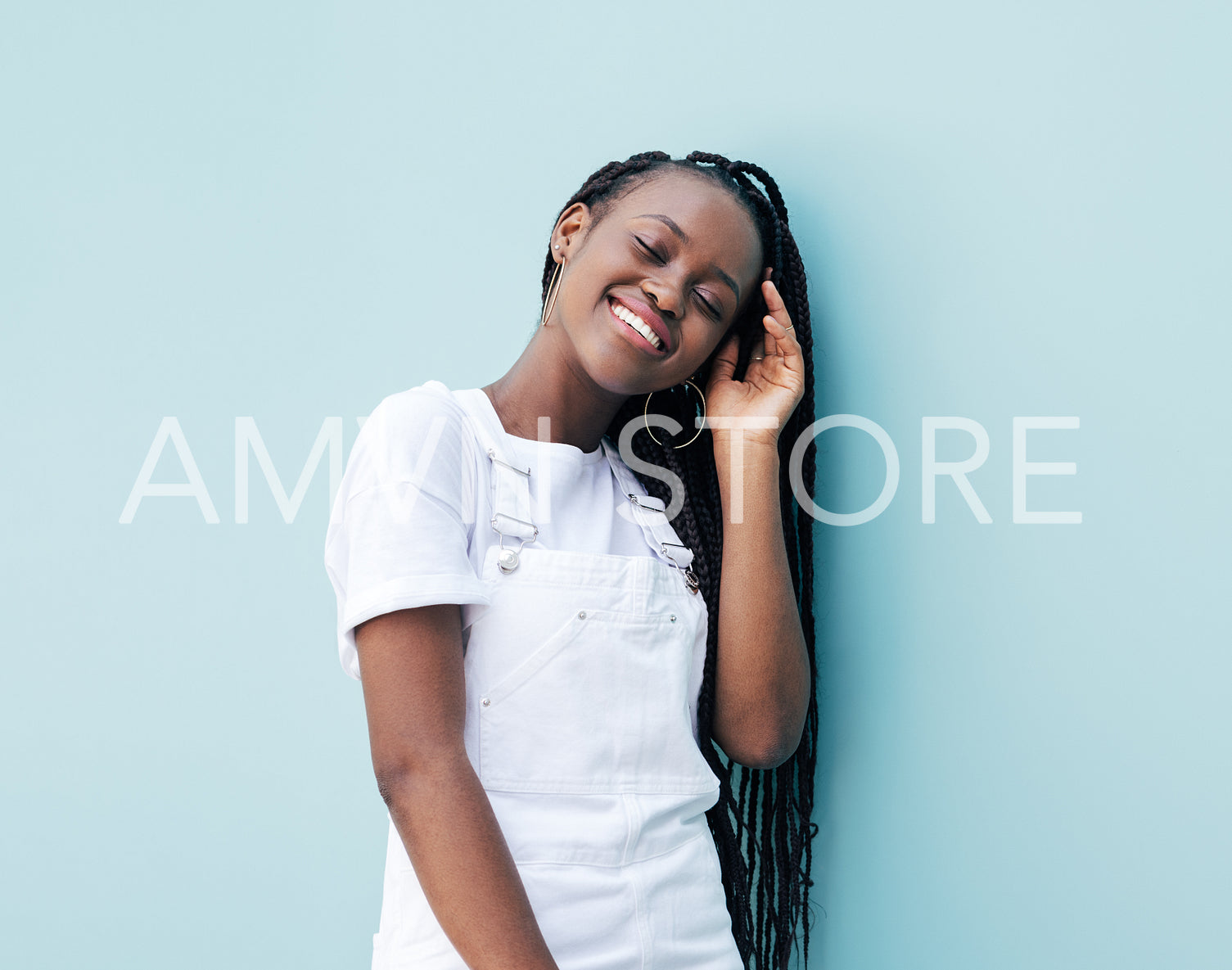 Cheerful female with braids wearing white clothes leaning on a blue wall with closed eyes