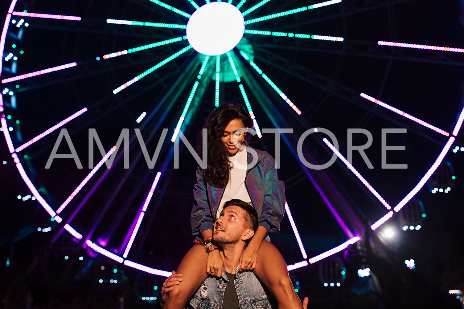 Smiling woman sitting on the shoulders of her boyfriend and looking at him. Young couple at night against ferris wheel with colorful lights.