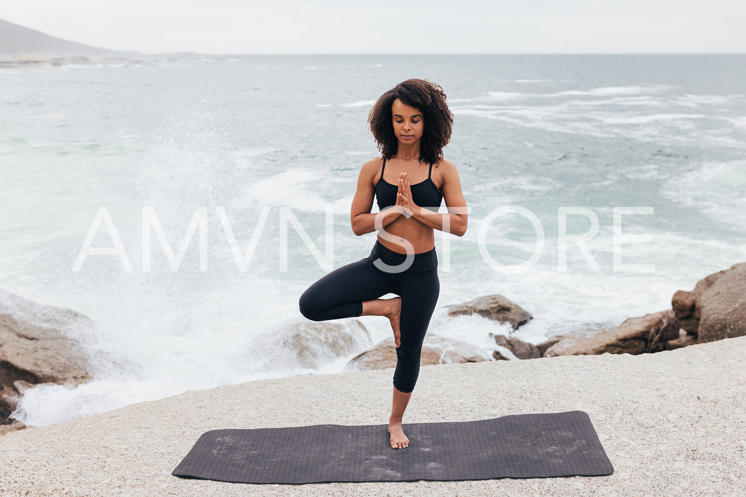 Woman with curly hair practicing Tree Pose by seaside. Young female with closed eyes doing Vrksasana at evening.