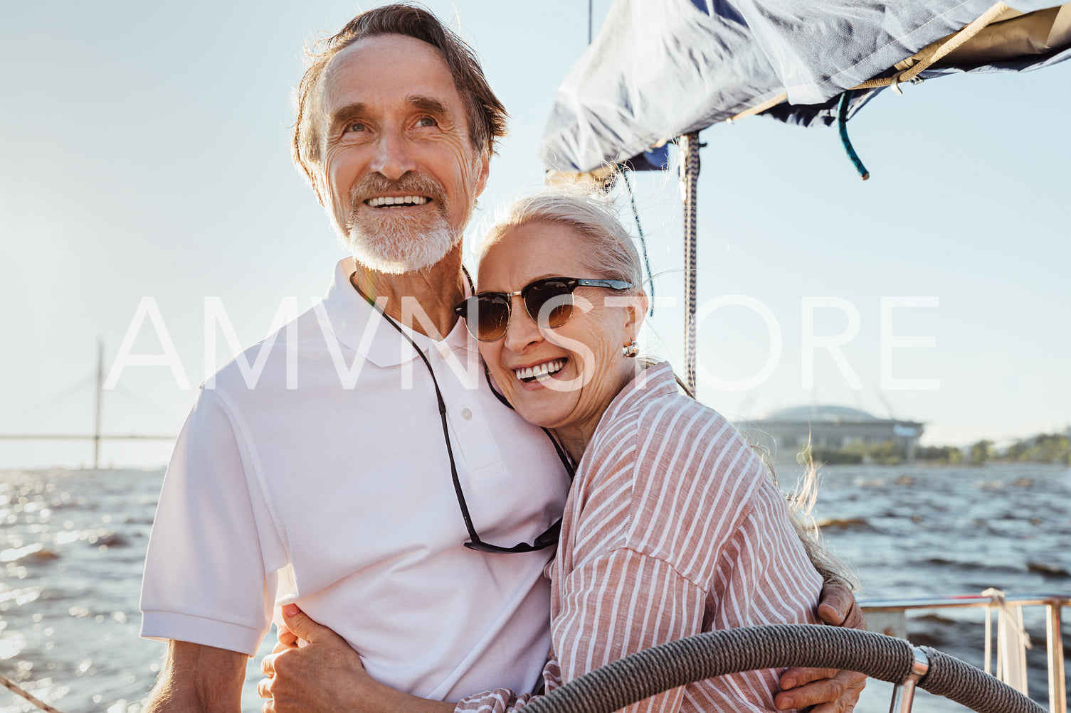 Cheerful senior couple standing at steering wheel on a yacht and hugging each other. Happy people enjoying sunny day on a boat.	