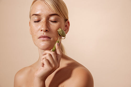 Blond woman with freckles using jade roller while standing with closed eyes against pastel background