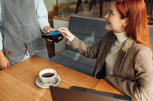 Woman paying by credit card with NFC in a coffeeshop. Female with ginger hair sitting at the table paying her bill.