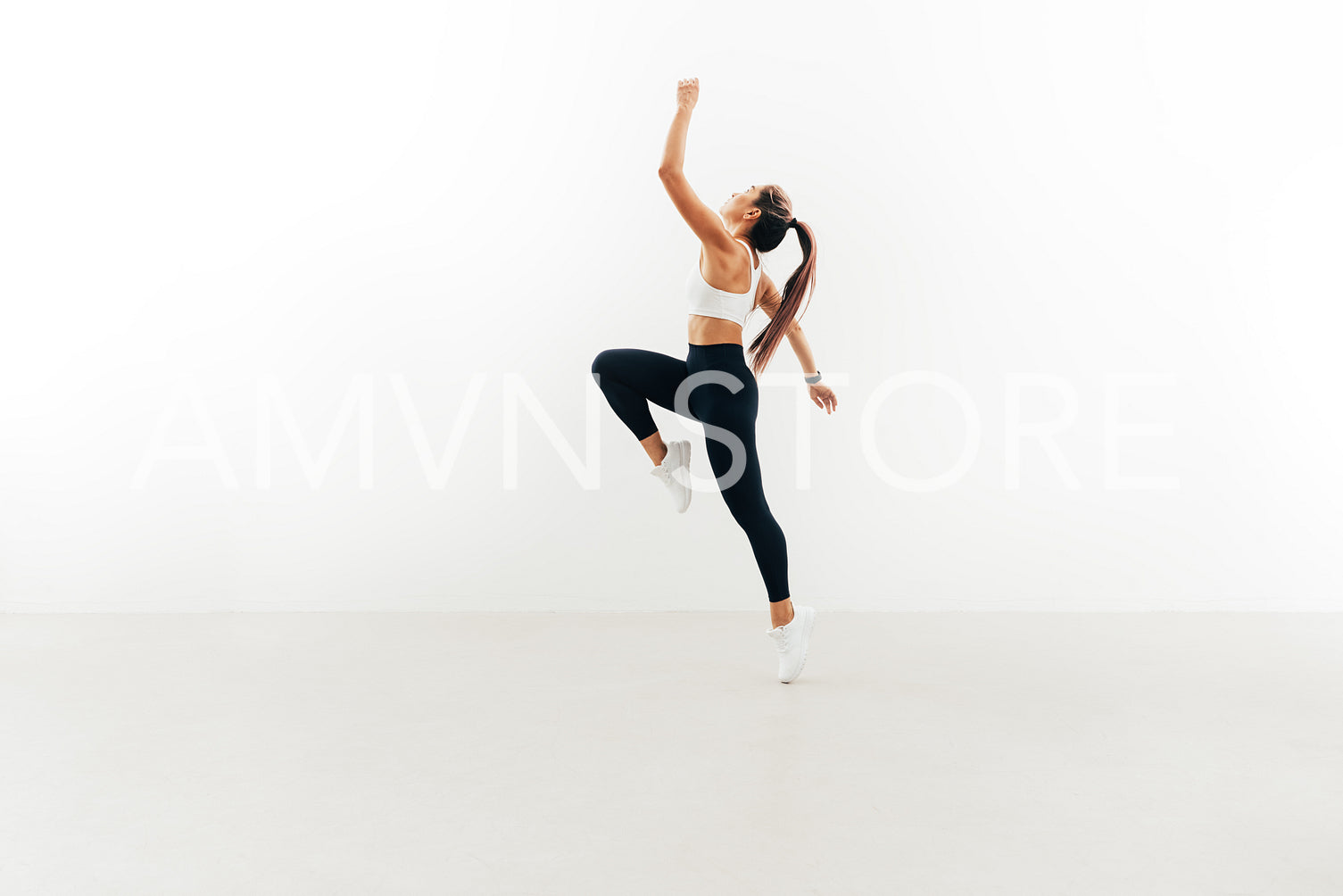 Side view of young female athlete jumping in the air raising hand up against white wall