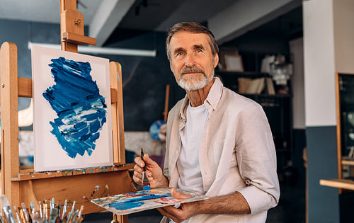 Portrait of a senior painter holding palette with a brush and looking away. Artist in his studio.