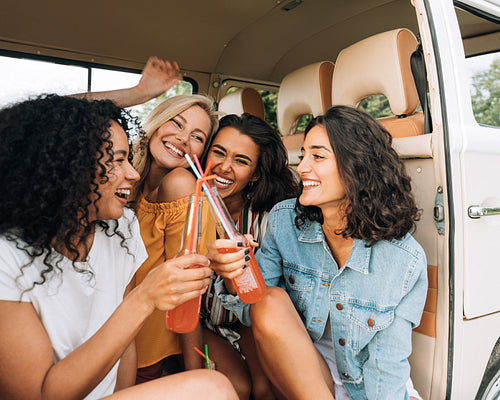 Happy women sitting together in van during road trip. Smiling females having fun and drinking cocktails in car.