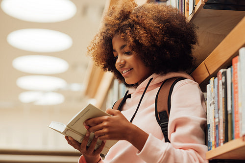 Girl in casuals with backpack leaning bookshelf in library. Student reading a book while standing in library.