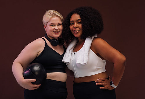 Two plus size females with fitness accessories standing together on brown background