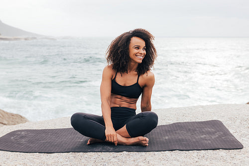 Smiling woman looking away while sitting on yoga mat by ocean. Female relaxing after yoga exercises.