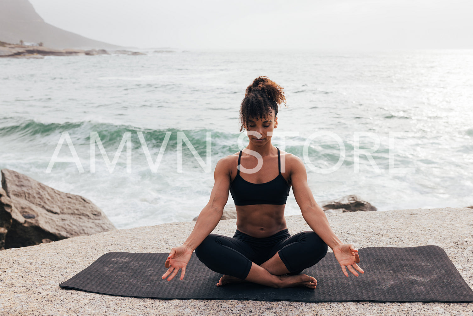 Woman with closed eyes sitting in lotus pose on mat by ocean. Female meditating at evening at seaside.