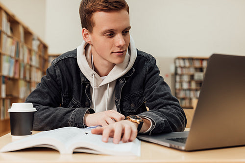 Male student sitting at desk looking at laptop. Young guy in library preparing exams.