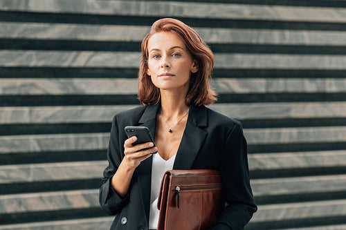 Middle-aged businesswoman holding a leather folder and smartphone and looking at camera while standing outdoors