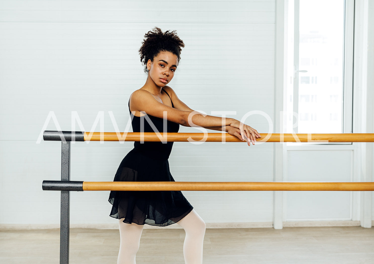 Young ballerina standing in dancing studio at barre and looking at camera	