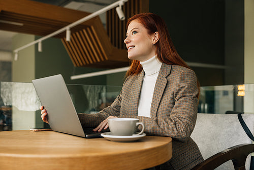 Woman entrepreneur sitting at a table with a laptop. Redhead female working remotely from a cafe.