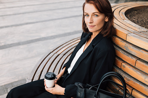 Middle-aged smiling businesswoman sitting on a bench outdoors. Female in black formal wear relaxing outdoors holding a coffee cup.