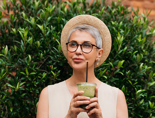 Stylish mature woman in a straw hat wearing eyeglasses holding a cocktail