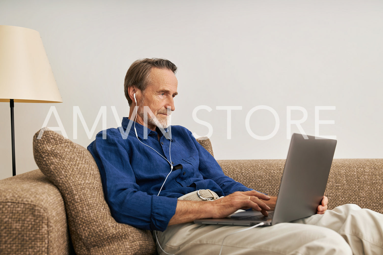 Side view of senior man wearing earphone typing on laptop. Elderly male sitting on sofa with laptop on his hips.	