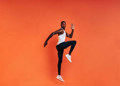 Young fit man doing exercises in studio. Male athlete jumping in the air over an orange background.