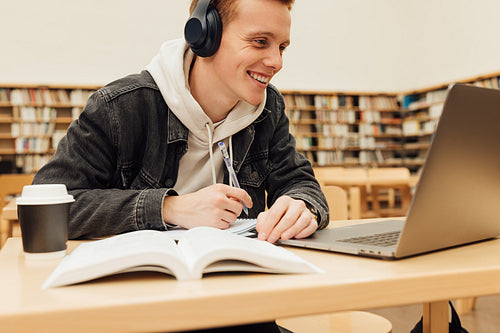 Young male student in wireless headphones preparing school assignment in library. Smiling man with laptop at college library.