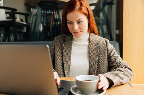 Businesswoman working in a coffee shop in the morning. Female with ginger hair in formal wear typing on laptop and drins coffee.