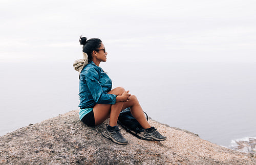 Woman hiker relaxing on a rock. Female mountain climber in sports clothes and sunglasses looking at the view.