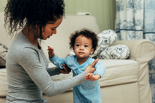 Mother sitting on the floor in room and teaching her baby son to walk