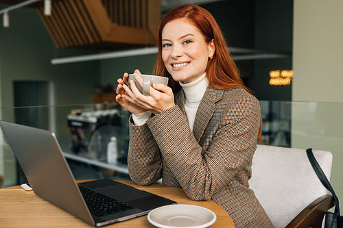 Smiling businesswoman holding a cup of coffee and looking at the camera. Female in business casual clothes sitting at a table in a cafe.