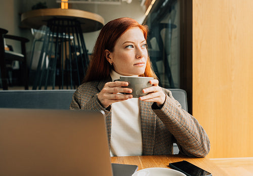 Thoughtful woman with ginger hair holding a cup of coffee sitting in a cafe looking at window