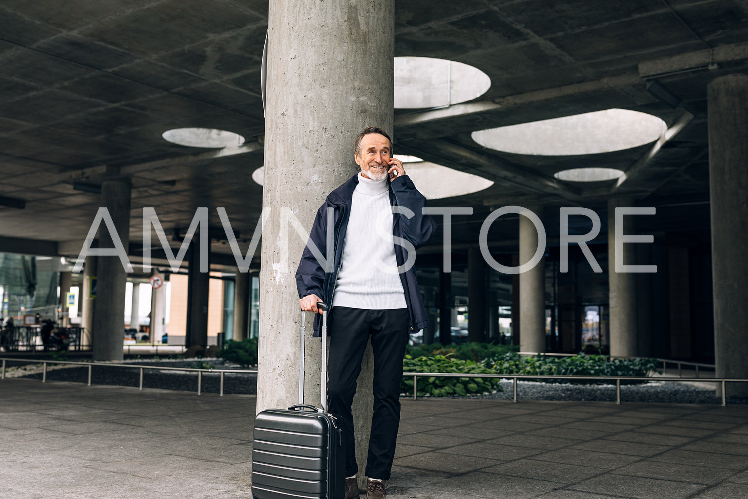 Smiling mature traveller standing outdoors with luggage and talking on cell phone