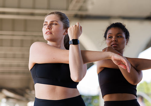 Two females stretch their hands while practicing under a bridge. Young plus-size woman doing warming-up exercises with her friend and looking away.