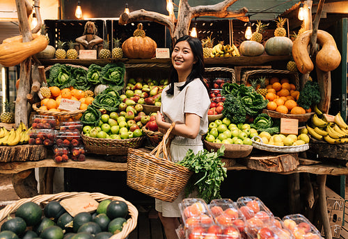 Side view of an Asian woman with a wicker shopping basket standing on a local outdoor market. Smiling female buying organic food.