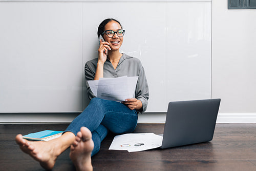 Smiling woman making a phone call while sitting on a floor with documents in hand